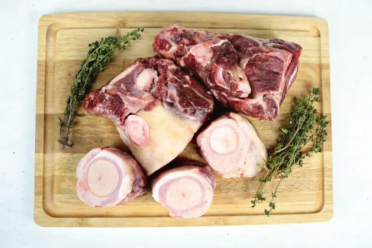 A wooden cutting board with raw beef soup bones surrounded by sprigs of fresh thyme.