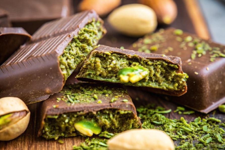 Close-up of kunafa chocolate bars filled with pistachio cream, surrounded by whole pistachios and sprinkled with crushed pistachio bits.