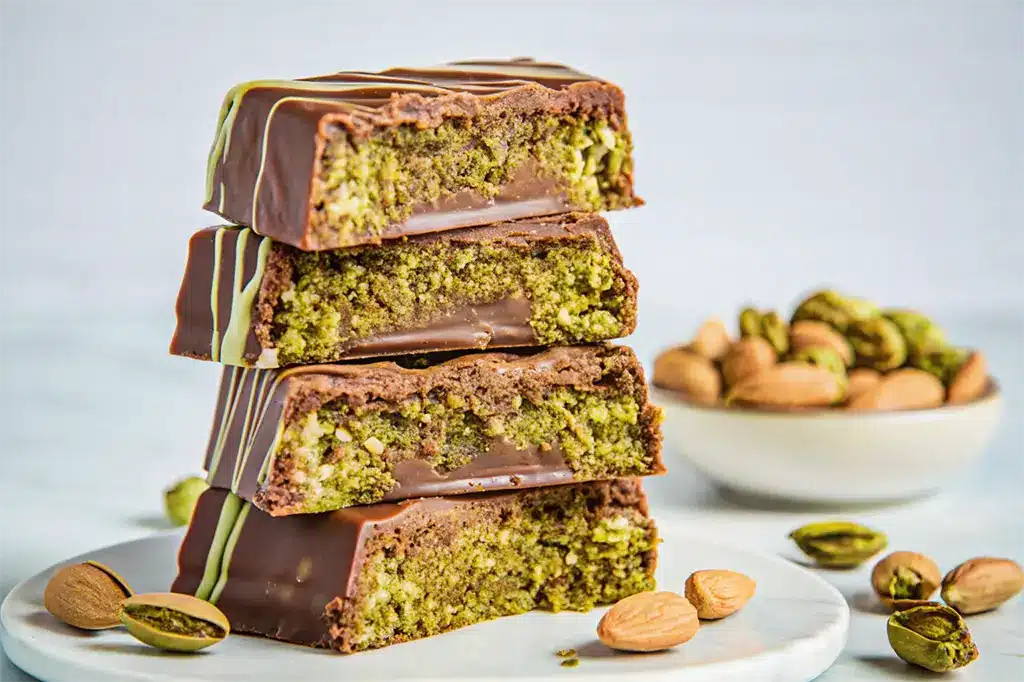 A stack of knafeh chocolate bars filled with pistachio cream and coated in rich chocolate, surrounded by whole pistachios and almonds.