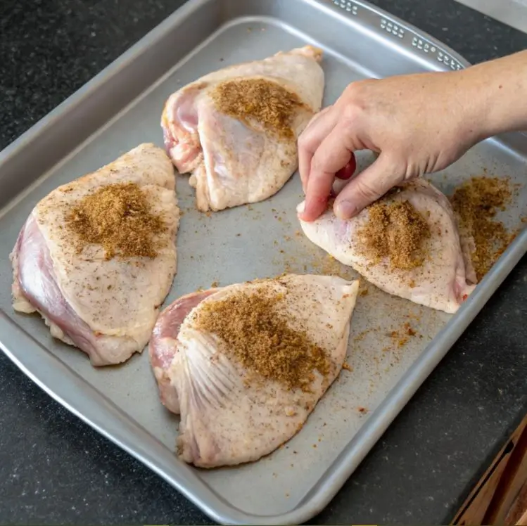 The image shows a baking tray with raw poultry pieces, likely turkey tails or chicken parts, being seasoned. A hand is seen sprinkling a brown seasoning mix onto the poultry. The tray is placed on a dark countertop, and the poultry is evenly spaced, ready for further preparation such as roasting or baking. The seasoning mix appears to be a blend of spices, possibly including paprika, garlic powder, onion powder, or other similar ingredients.