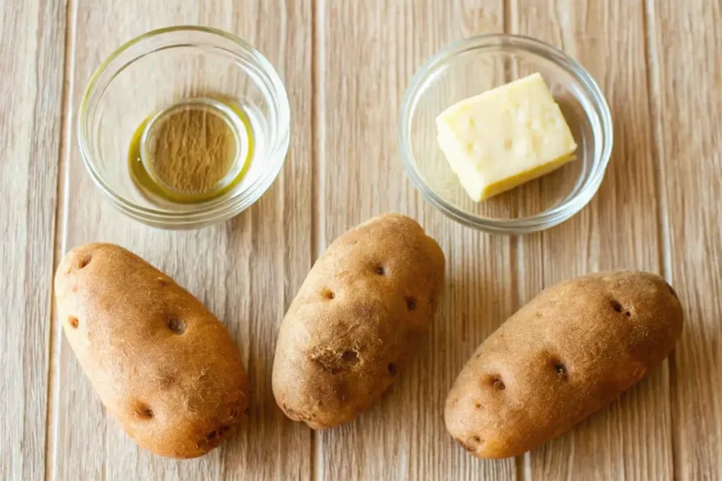 Three raw potatoes placed on a wooden surface, accompanied by small glass bowls containing butter and olive oil.