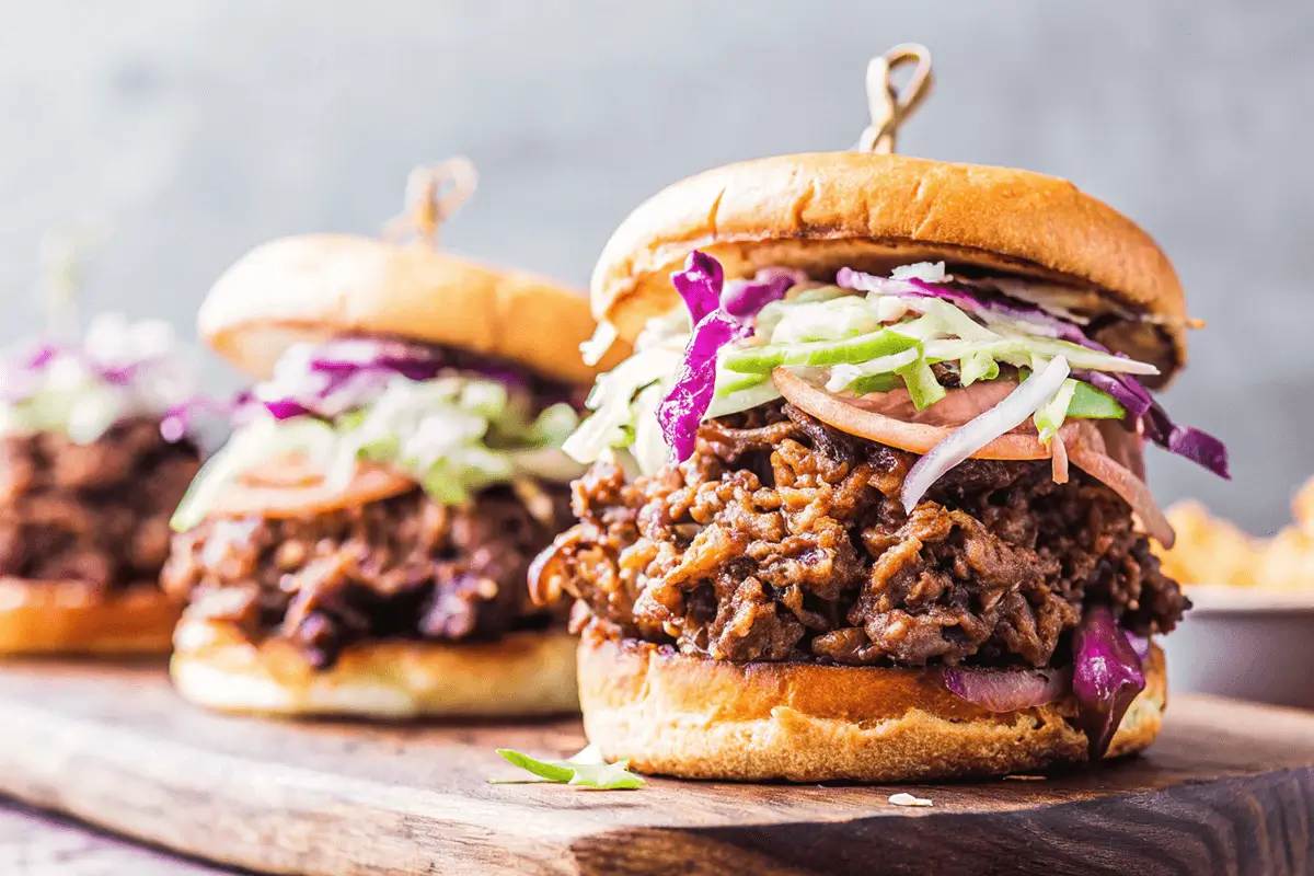 what goes with brisket sandwiches? A close-up of pulled beef barbecue sandwiches on a wooden board. The sandwiches are topped with shredded coleslaw and served on soft brioche buns.