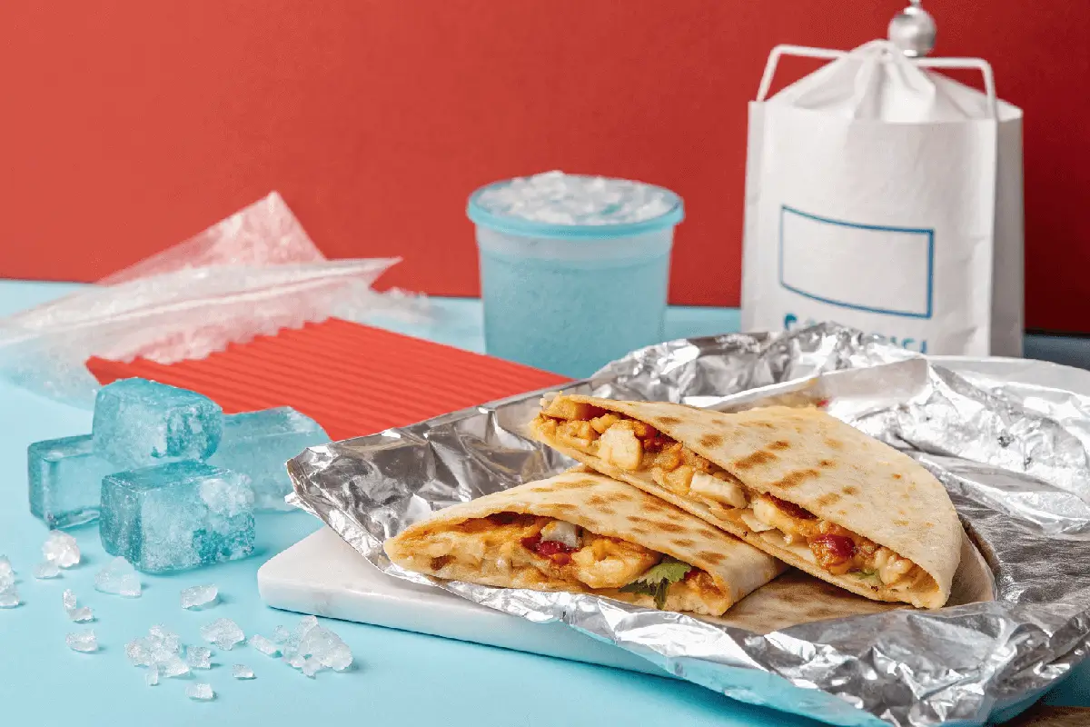 Can I Freeze Stuffed Quesadillas? A stuffed quesadilla is folded in half and cut into halves, placed on a foil wrapper, on a light blue surface with ice cubes nearby. A blue slush drink, a white takeaway bag, and a red background complete the scene