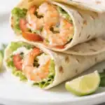 Close-up of a fresh shrimp wrap recipe with juicy shrimp, lettuce, tomato, and creamy sauce inside a tortilla, served with lime wedges on a white plate.