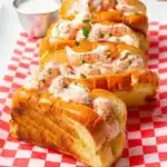 Four golden langostino lobster tails served in toasted buns on a red-and-white checkered paper, garnished with fresh chives and a side of creamy sauce.
