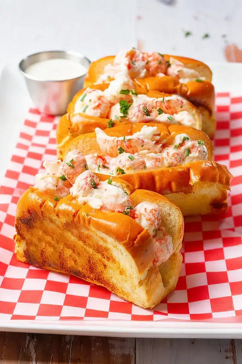 Four golden langostino lobster tails served in toasted buns on a red-and-white checkered paper, garnished with fresh chives and a side of creamy sauce.