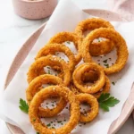 Crispy keto onion rings served on a pink speckled platter with parchment paper and a side of red dipping sauce, garnished with fresh parsley.