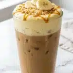 Tall glass of iced coffee topped with thick banana pudding cold foam, caramel drizzle, crushed vanilla wafers, and fresh banana slices.