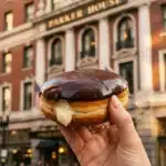 Hand holding a chocolate frosted Boston cream donut with custard filling in front of the historic Parker House Hotel in Boston.