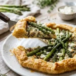 A slice of rustic asparagus galette with golden puff pastry, roasted green asparagus, and crumbled cheese is being lifted from a white plate.
